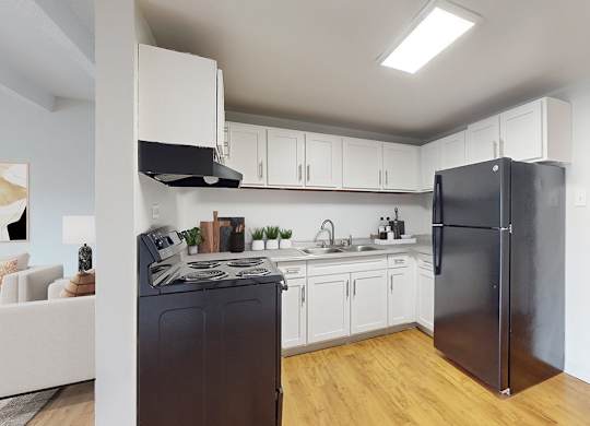 A black refrigerator stands in a kitchen with white cabinets and a wooden floor.