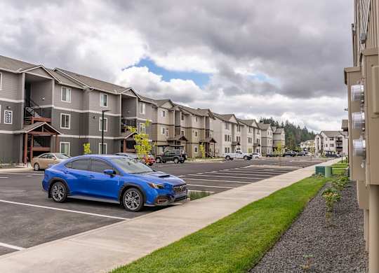 A blue car is parked in a parking lot in front of apartment buildings at Marcola Apartment Homes, Forest Grove, OR