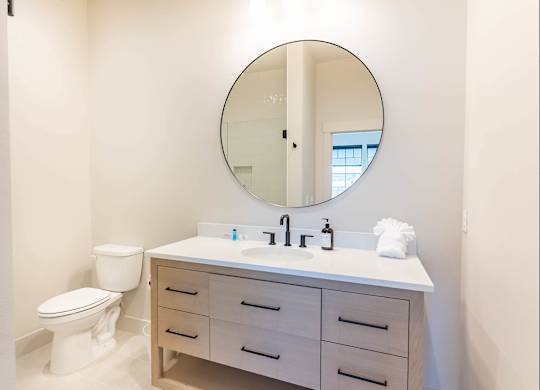 A white bathroom with a round mirror and a white toilet at Westgate Apartment Homes, Pendleton, OR