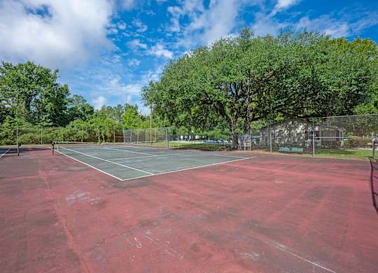 A red tennis court surrounded by trees and a fence at Sawbranch Apartments, Summerville