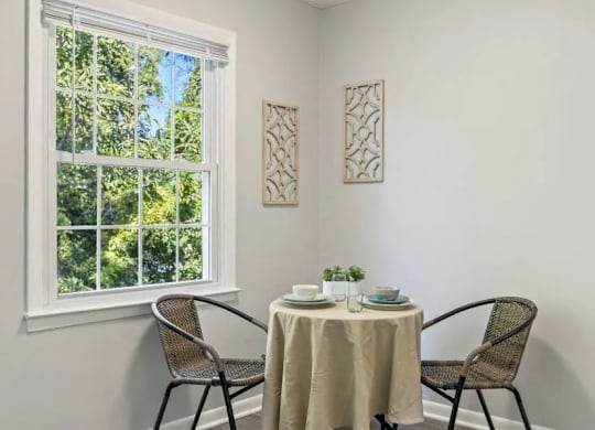 A table with a white tablecloth and two chairs is set up in front of a window at The Preserve at Pine Valley Apartments, North Carolina 28412