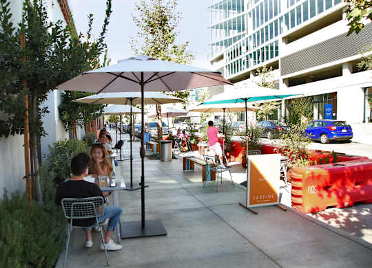 A man and woman are sitting at a table under an umbrella in a courtyard.at Prizma, Los Angeles, 90038  
