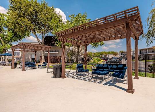 A wooden pergola with blue cushioned seating is in the foreground of a sunny park.