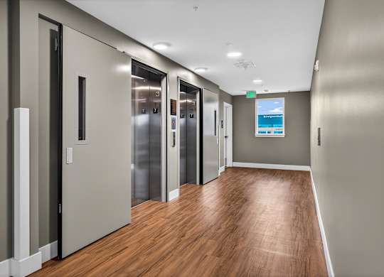 A long hallway with wood floors and a row of elevators.