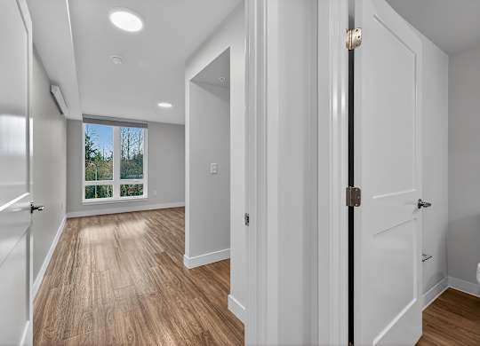 A hallway with white walls and wooden floors leading to a bright window.