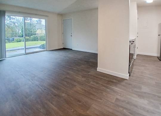 A spacious room with wooden flooring and a view of a green outdoor area through the windows.
