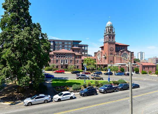 A church with a clock tower is in the background of a street with cars parked on the side.