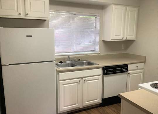 A white refrigerator stands in a kitchen with white cabinets and a window with blinds.