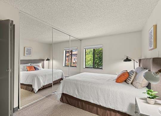 Apartments Near St. Joseph Medical Center, Tacoma, WA - The Orion - Bright Bedroom With Mirrored Closet Doors, Neutral Carpet, White Bedding, Bedside Lamps, and Window.