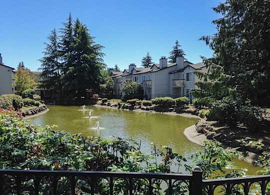 A pond with a fountain in the middle of a garden with a house in the background.