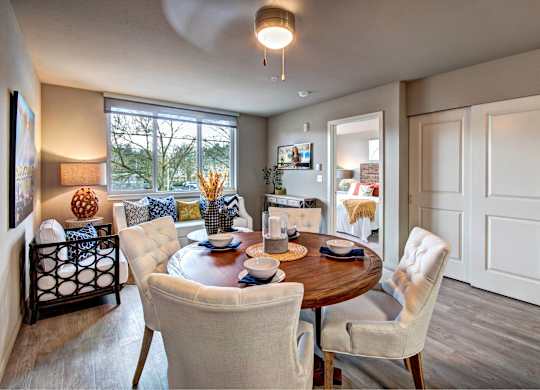 A dining room with a table set for two and a view of trees outside the window.