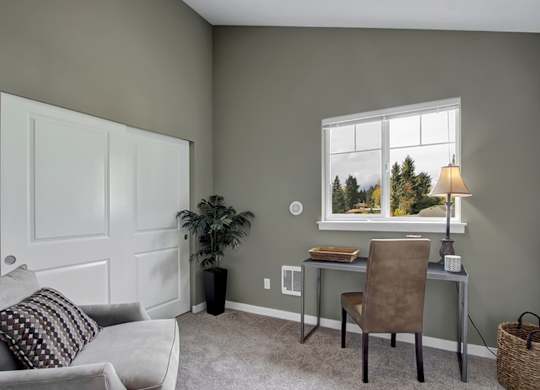 A room with a grey couch, a brown chair, a desk with a lamp and a window showing trees outside.