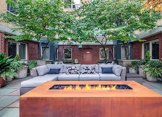 A fire pit in the middle of a patio surrounded by a white couch and potted plants.
