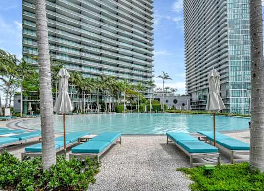 A pool surrounded by palm trees and benches with a building in the background.