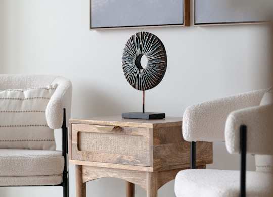 A room with a white chair, a wooden table with a fan on it, and a painting on the wall.