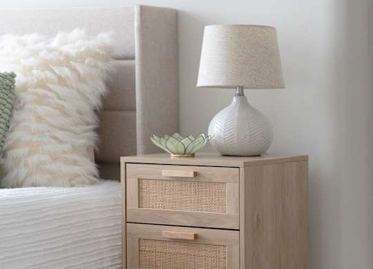 A white lamp with a green plant on top of a wooden nightstand.