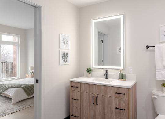 A bathroom with a white toilet, a sink with a mirror above it, and a bedroom with a bed and a window.
