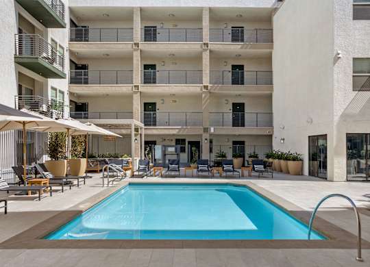 A large swimming pool is surrounded by lounge chairs and umbrellas in front of a multi-story apartment building.