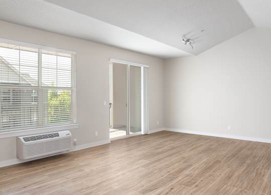 A room with a slanted ceiling, a window with blinds, a wall-mounted air conditioning unit, and a wooden floor.