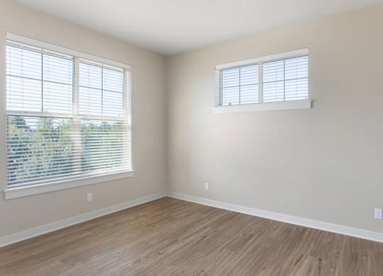 A room with wooden flooring and two windows with blinds.