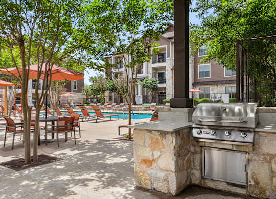 A stone BBQ grill is in the middle of a patio with chairs and tables.