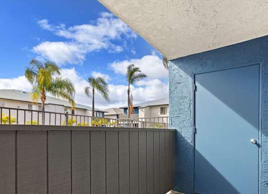 A blue door is on the right side of a wall with a balcony in the background.