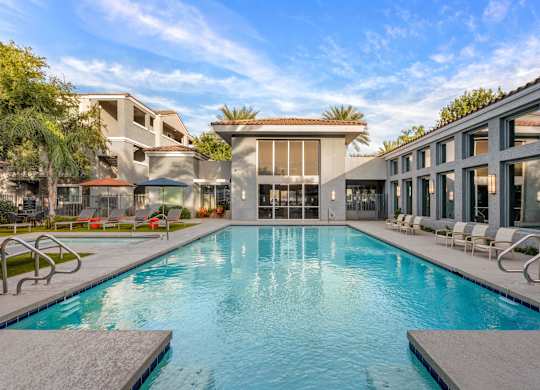 A large swimming pool in front of a building with lounge chairs around it.