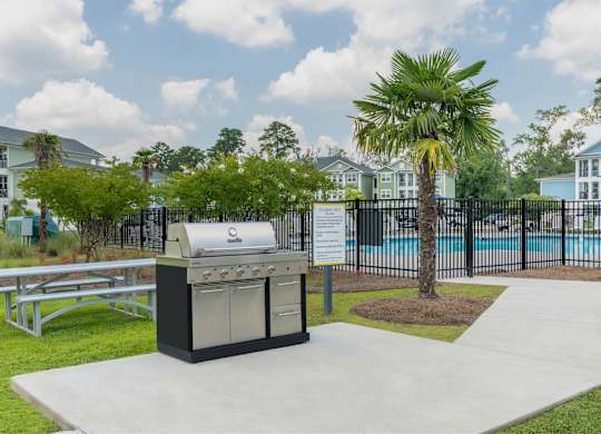 A BBQ grill is in the middle of a concrete area with a pool and palm tree in the background.