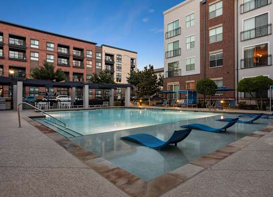 A swimming pool surrounded by a poolside lounge area and apartment buildings.