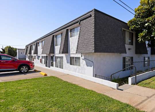 A red car is parked in front of a white building with a black roof.