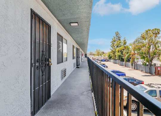 A long, narrow balcony with a metal railing and a white wall with a black gate.