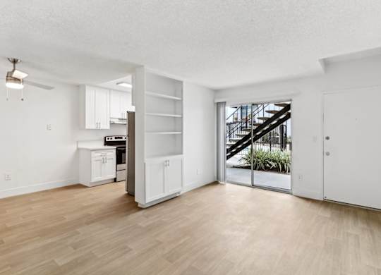 A room with a wooden floor and white walls, with a kitchen area and a staircase visible through the glass door.