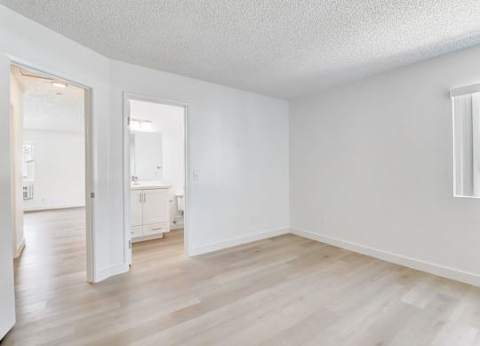 A room with white walls and wooden flooring, with a window showing a building outside.
