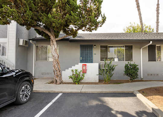 A black car is parked in front of a two-story house with a tree in front of it.