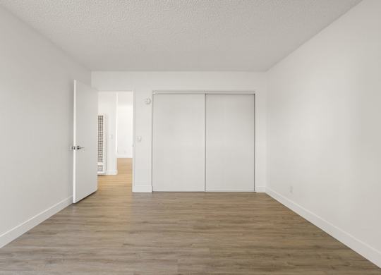 A room with white walls and wooden flooring with a door on the left and a closet on the right.