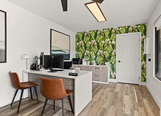 A room with a desk and chairs in front of a window with a view of a green plant.