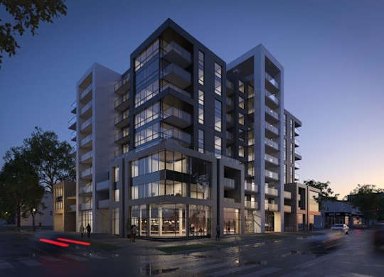 A modern multi-story building with a glass facade is illuminated from within, with a street in front of it.