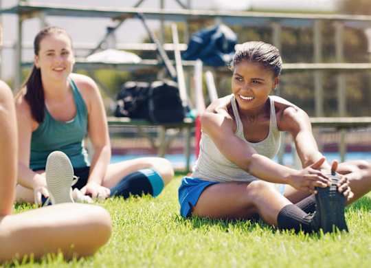 Three women are sitting on the grass, one of them is tying her shoelaces.