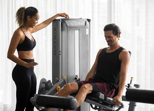A woman is adjusting the settings on a gym machine while a man sits on a rowing machine. at Palma, Doral, FL
