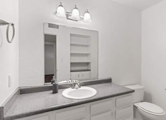 A bathroom with a sink, mirror, toilet and a towel ring on the wall.