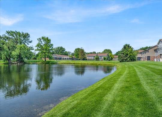 A serene landscape with a lake, grass, and residential buildings.