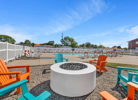 A white fire pit sits in the middle of a gravel area with orange and blue chairs surrounding it.
