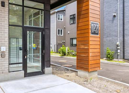 The image shows a modern building with a glass door and a wooden column with the number 177 on it.