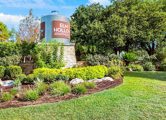 A sign for Elm Hollow Apartments is displayed on a water tower in a landscaped area.