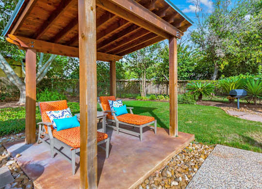 A wooden gazebo with orange cushions and a bench is surrounded by a garden.