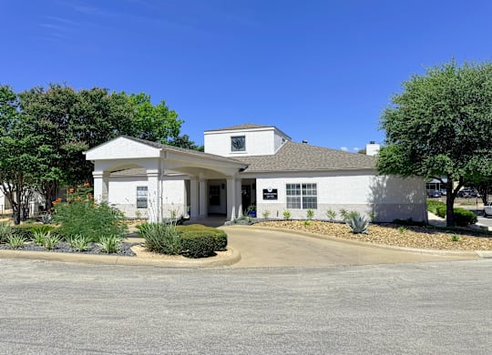 A white building with a black signboard is surrounded by green plants and trees.