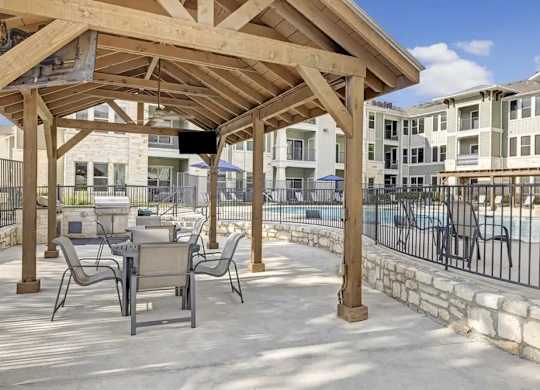 A wooden pergola is over a patio with chairs and a pool in the background.