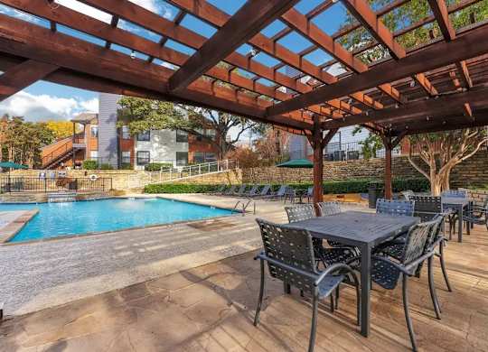 A wooden pergola over a table with chairs and a pool in the background.