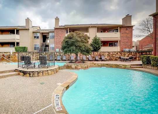 A swimming pool surrounded by a stone wall and a patio area with chairs and a table.