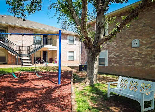 A playground with a swing set and a bench is in front of a brick building with the number 27 on it.